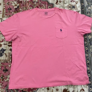 Rosa t-shirt från Polo Ralph Lauren - Snygg rosa t-shirt från Polo Ralph Lauren med klassisk passform. T-shirten har korta ärmar, rund halsringning och en bröstficka med den ikoniska blå broderade loggan.