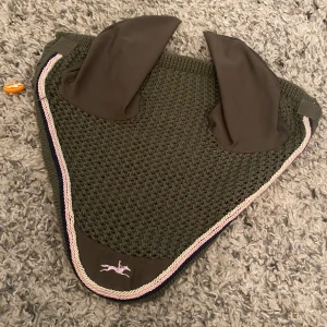 Olivgrön ridhuva Schockemöhle Sports - Aldrig använd! Snygg olivgrön ridhuva från Schockemöhle Sports i storlek Full. Stickad kropp med släta öron i syntetmaterial, kantad med rosa, vit och marinblå snodd. Broderad hästlogga framtill. Perfekt för dig som vill ge din häst en cool look på träning eller tävling.
