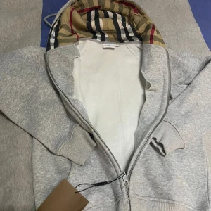Grå Burberry hoodie med klassisk huva - Grå hoodie från Burberry med dragkedja och ribbade muddar. Huvan har det ikoniska Burberry-rutiga fodret i beige, svart, vitt och rött. Tröjan har snörning i huvan med snygga metalldetaljer. Perfekt för dig som vill ha en stilren och lyxig look.