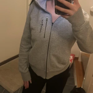 Grå hoodie från Sail Racing med huva - Grå hoodie från Sail Racing med dragkedja framtill och en extra zip-detalj på bröstet. Mjuk insida i fleece och klassisk huva. Perfekt för chill dagar och har en clean look med logga på bröstet. Passar dig som gillar sportig och enkel stil.