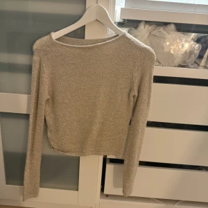 Glittrig beige stickad topp - Säljer en glittrig beige stickad topp med lång ärm och rund hals. Toppen har en lätt croppad passform och är perfekt för dig som vill sticka ut lite extra. Materialet är mjukt och skimrar snyggt i ljuset.
