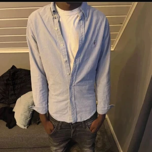 Blå skjorta från Ralph Lauren slim fit - Snygg ljusblå skjorta från Ralph Lauren i slim fit-modell. Klassisk design med knappar framtill och broderad logga på bröstet. Skjortan är långärmad och tillverkad i mjuk bomull, perfekt för en clean och stilren look.