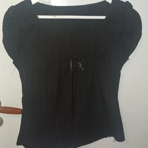 Svart blus med knyt och puffärm - Svart blus med subtilt randigt tyg, v-ringning och söt knytning framtill. Puffiga korta ärmar ger en trendig siluett. Inte stretch, har ett håll vid armen men kan nog fixas :) vet inte riktigt storleken för det står inget o den e asiatisk storlek men gissar typ M kanske? 