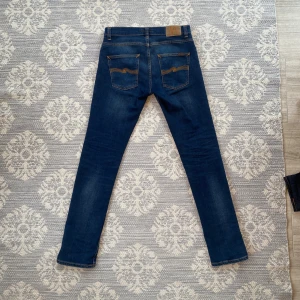 Nudie Jeans Grim Tim Indigo Myth - Snygga mörkblå jeans från Nudie Jeans, modell Grim Tim Indigo Myth. Klassisk femficksdesign med kontrastsömmar och ikonisk broderad bakficka. Tillverkade i stretchig bomull med lite elastan för extra komfort. Perfekta för dig som gillar en smal passform.