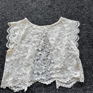 Vit spetsig bolerotopp med blommönster - Superfin vit bolerotopp i spets med blommigt mönster och vågiga kanter. Toppen är ärmlös och har öppen rygg, perfekt att slänga över ett linne eller klänning för en extra touch. (Pris kan diskuteras)