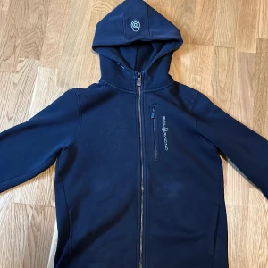 Svart Sail Racing zip - Snygg svart hoodie från Sail Racing med hel dragkedja och bröstficka med logga. Tröjan har huva och är tillverkad i en mix av bomull och polyester. Perfekt för dig som gillar sportig och stilren look. Den ser ut att vara marinblå men den är svart. Ny pris 1000kr