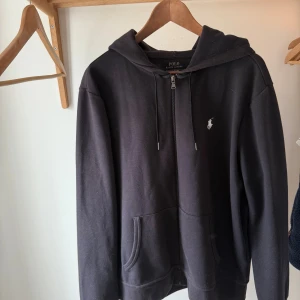 Svart hoodie från Polo Ralph Lauren - Svart hoodie från Polo Ralph Lauren med dragkedja framtill, huva med snörning och klassisk vit logga på bröstet. Tröjan har känguruficka och är tillverkad i mjuk bomull. Perfekt för chill dagar eller när du vill ha en clean streetstyle.