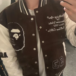 Brun och vit varsityjacka BAPE - Säljer en brun och vit varsityjacka från BAPE med broderade detaljer, stora loggor och text på både fram- och baksida. Jackan har vita ärmar, bruna muddar med ränder och knappar framtill. Höger ficka är sönder och några andra skavanker som man egentligen inte tänker på. Därmed det billiga priset. Pris kan diskuteras vid snabb affär.