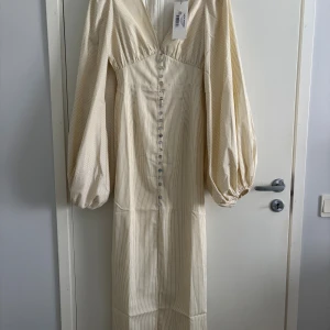Randig långklänning från Adoore - Säljer en Positano Maxi Dress från Adoore i ljusgul och med smala ränder. Klänningen har v-ringning, knappar framtill och puffiga långärmar med resår vid ärmslut. Aldrig använd och med prislapp kvar💕🌼