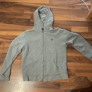 Grå hoodie från Polo Ralph Lauren - Snygg grå hoodie från Polo Ralph Lauren med dragkedja och huva. Klassisk design med broderad logga på bröstet och två fickor framtill. Tillverkad i mjuk bomullsmix som är perfekt för kyliga dagar.