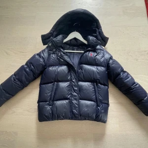 Mörkblå pufferjacka från Polo Ralph Lauren - Säljer en mörkblå pufferjacka från Polo Ralph Lauren med avtagbar huva och röd broderad logga på bröstet. Jackan har dragkedja framtill och är vadderad för extra värme. Perfekt för kalla dagar och riktigt snygg stockholmstill.                                Jackan är köpt från kidsbrandstore. Den passar för 12-14 åringar. Men också 15 åringar. Jackan har jag andvänt en vinter säsong!🥰Det är bara att skriva om ni har mer frågor