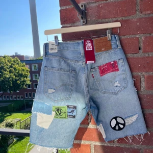 Levi's 501 patchade jeansshorts - Helt nya och inköpta på en Levis butik i New York! Unika ljusblå jeansshorts från Levi's 501 med coola patchar, broderade märken och slitna detaljer. Shortsen har hög midja, råa kanter och flera färgglada tygbitar och badges, bland annat peace-symbol och bandana-tyg. Perfekt för dig som vill sticka ut i sommar.