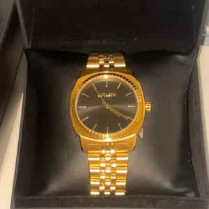 Guldig klocka från Lucleon - Stilren klocka från Lucleon med guldig länk och svart urtavla. Klockan har en klassisk rund form och minimalistisk design med tunna indexmarkeringar. Perfekt accessoar för att lyfta din outfit.