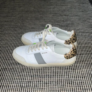 Arigato sneakers med leoparddetalj - Snygga vita sneakers från Arigato med gråa ränder på sidorna och cool leopardmönstrad detalj på hälen. Skorna har snörning och platt sula, tillverkade i skinn med inslag av syntet. Perfekta för dig som vill sticka ut lite extra.