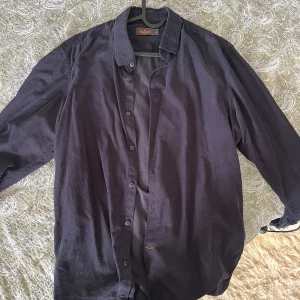 Svart manchesterskjorta från Morris - Säljer en svart långärmad skjorta från Morris i manchestertyg. Skjortan har klassisk krage, knäppning framtill och diskret broderad logga på bröstet. Perfekt för dig som gillar stilrena och tidlösa plagg. Nypris 1200