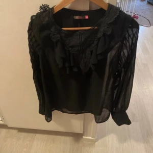 Svart blus med spets och volang - Säljer en svart blus i transparent tyg med långa ärmar och snygga detaljer. Blusen har spets och volang vid halsringningen, samt dekorativa knappar vid ärmsluten. Ärmarna har ett flätat mönster som ger extra edge till looken.