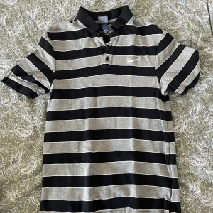 Randig pikétröja från Nike - Snygg pikétröja från Nike med breda svarta och grå ränder, klassisk krage och tre knappar framtill. Tröjan har korta ärmar och är tillverkad i mjuk bomull. Perfekt för en sportig och avslappnad stil.