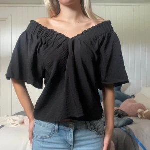 Volangblus - Säljer en svart offshoulder blus med bred resår upptill som ger en snygg rynkad effekt. Blusen har vida, korta ärmar och en luftig passform. Perfekt att styla med jeans eller kjol för en chill look. Fast pris!