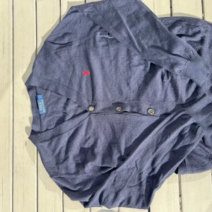 Mörkblå kofta från Polo Ralph Lauren - Klassisk mörkblå kofta från Polo Ralph Lauren med knappar framtill och röd broderad logga på bröstet. Långärmad och tillverkad i mjuk bomull, perfekt för lager-på-lager. Enkel och stilren design som funkar till det mesta.
