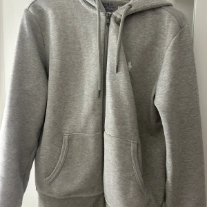 Grå hoodie från Polo Ralph Lauren - Säljer en stilren grå hoodie från Polo Ralph Lauren med dragkedja framtill och klassisk huva. Tröjan har två fickor och diskret logga på bröstet. Perfekt för en chill och avslappnad look. Materialet är mjukt och skönt, troligtvis bomullsmix.