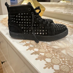 Svarta sneakers med nitar - Säljer ett par svarta high-top sneakers med ikoniska röda sulor. Skorna är täckta med svarta nitar och har snörning framtill. Materialet är mocka och detaljerna ger en edgy vibe. Perfekt för dig som vill sticka ut med din stil.