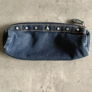 Jeans pennfodral med nitar och dragkedja - Pennfodral i mörkblå jeans med coola silverfärgade nitar längs ovansidan. Stängs med dragkedja och har en detalj med knapp i ena hörnet. Stilren och enkel design som passar till det mesta.