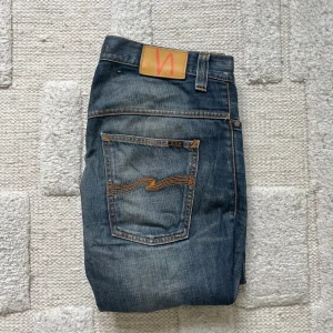 Blå jeans från Nudie Jeans Co - Säljer ett par klassiska blå jeans från Nudie Jeans Co med snyggt slitna detaljer och kontrastsömmar. Jeansen har fem fickor, orange broderi på bakfickorna och normal passform. Tillverkade i bomull och har en rak siluett.