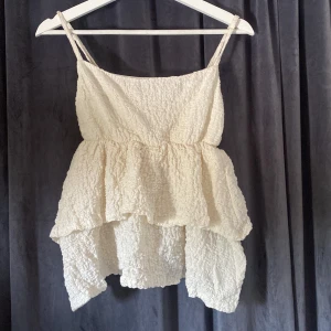 Beige volangtop från Gina Tricot - Supersöt beige linnetopp från Gina Tricot med volanger och smala axelband. Toppen har en crinklad struktur och är i ett lätt, syntetiskt material. Perfekt för sommarens alla tillfällen när du vill känna dig extra fin.