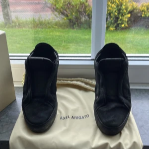 Svarta sneakers från Axel Arigato - Stilrena svarta sneakers från Axel Arigato i mocka med minimalistisk design och platt sula. Skorna har rund tå och diskret branding, perfekta för dig som gillar en clean look. Kommer med originalpåse och box.tyvär har arigato texten nästan åkt bort. Pris kan sänkas vid snabb affär 