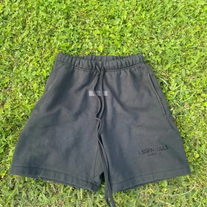 Svarta shorts Essentials Fear of God - Svarta shorts från Essentials Fear of God med elastisk midja och långa snören framtill. Klassisk loose fit och diskret logga på vänster ben. Perfekta för chill dagar eller streetwear. Tillverkade i mjuk bomull för maximal komfort.