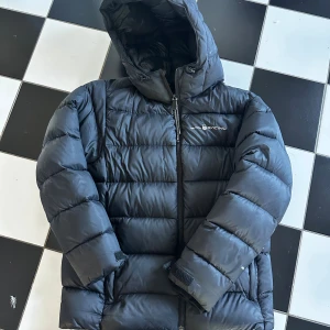 Svart pufferjacka från Sail Racing - Svart pufferjacka från Sail Racing med huva och dragkedja framtill. Jackan har en quiltad design och logga på bröstet. Perfekt för kalla dagar och har en riktigt skön, vadderad känsla. Snygg och enkel att matcha med det mesta.