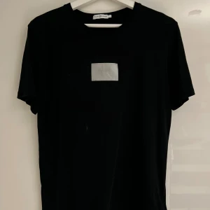 Svart t-shirt från Calvin Klein Jeans - Svart t-shirt från Calvin Klein Jeans med en vit patch och diskret CK-logga på bröstet. Klassisk rund hals och korta ärmar. Mjuk och skön bomullskvalitet som passar perfekt till jeans eller shorts.