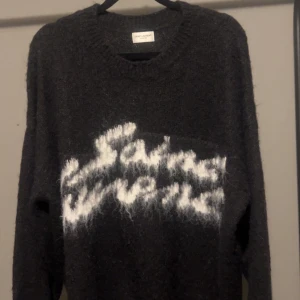 Svart Saint Laurent stickad tröja - Säljer en svart stickad tröja från Saint Laurent med vit, fluffig logga framtill. Tröjan har lång ärm och en avslappnad passform. Perfekt för dig som vill ha en statement piece med lyxig känsla och cool design.