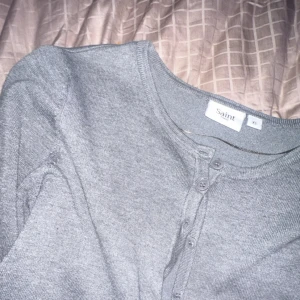  kofta med knappar - Grå, klassisk kofta från Saint i storlek XS. Koftan har rund halsringning, långa ärmar och knäppning framtill med matchande knappar. Tillverkad i mjuk bomull och passar perfekt för lager-på-lager-stil.