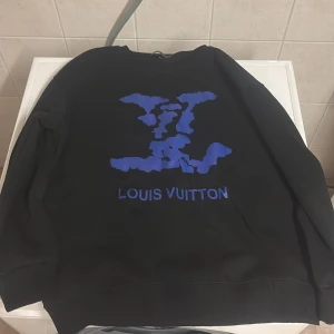 Svart sweatshirt Louis Vuitton blå print - Svart sweatshirt från Louis Vuitton med ett stort blått LV-print på framsidan. Tröjan har rund hals och ribbade muddar vid ärmslut och nederkant. Materialet känns mjukt och skönt, perfekt för dig som gillar exklusiva streetwear-vibbar.