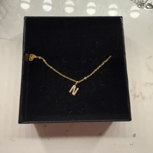 Guldhalsband med bokstav N i cubic zirconia - Snyggt halsband i guldpläterat 925 silver med en glittrig bokstav N som hänge, dekorerad med små cubic zirconia-stenar. Kedjan är tunn och elegant, perfekt för att ge din look en personlig touch. Märkt med S925 på låset.
