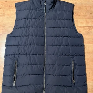 Marinblå dunväst från Bondelid - Svart quiltad dunväst från Bondelid med ståkrage och två fickor med dragkedja. Västen har en stilren look med horisontella sömmar och hel dragkedja framtill. Perfekt för lager-på-lager och enkel att matcha med olika outfits.