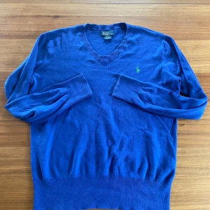 Blå v-ringad tröja Polo Ralph Lauren - Snygg blå v-ringad tröja från Polo Ralph Lauren i storlek XL. Tröjan har lång ärm och är dekorerad med det klassiska gröna Polo-logot på bröstet. Mjuk och tunn stickad känsla, perfekt för dig som gillar stilrena och tidlösa plagg.