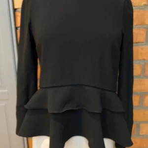 Svart volangblus från Zara Woman - Säljer en svart blus från Zara Woman i storlek S. Blusen har långa ärmar med två knappar vid manschetten, volangdetaljer nedtill och en stilren, enkel design. Materialet är mjukt och lätt, perfekt för dig som gillar en chic look med extra volym.