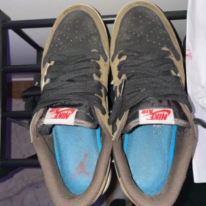 Nike Air Jordan 1 Low Travis Scott - Nike Air Jordan 1 Low Travis Scott med bruna och svarta paneler i mocka och skinn, vit omvänd swoosh och röda detaljer. Skorna har svarta snören, blå innersula med Jumpman-logga och klassisk Jordan 1 siluett. Perfekt för dig som gillar streetwear och unika sneakers.