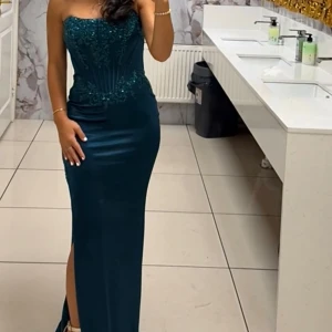Mörkgrön långklänning med paljetter - Säljer en elegant mörkgrön långklänning med figurnära passform. Klänningen är axelbandslös och har vackra paljettbroderier upptill samt en slits på sidan. Perfekt för bal eller fest, ger en lyxig känsla och snygg siluett.