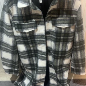 Rutig overshirt i ull från H&M - Snygg rutig overshirt från H&M i mjuk ullmix. Jackan har svart och gråa rutor på vit botten, klassisk krage, två bröstfickor med lock och knappar framtill. Perfekt för lager-på-lager och chill stil.