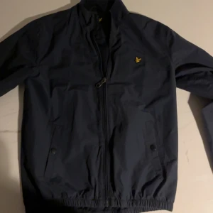 Marinblå vindjacka från Lyle & Scott i mycket bra skick  - Snygg marinblå vindjacka från Lyle & Scott med gul broderad logga på bröstet. Jackan har dragkedja framtill, två fickor med knappstängning och ribbad krage. Perfekt för dig som vill ha en stilren och sportig look.