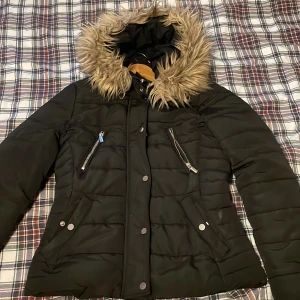 Svart pufferjacka med fuskpäls - Svart pufferjacka med snygg fuskpäls på huvan och quiltad design. Jackan har dragkedjor på bröstfickorna, tryckknappar framtill och en figurnära passform med markerad midja. Perfekt för kalla dagar och har en modern look.