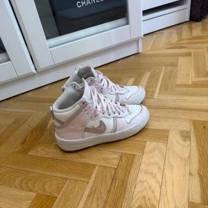 Nike Dunk High rosa/vit strl 37.5 - Säljer ett par Nike Dunk High sneakers i ljusrosa och vitt med klassisk snörning och rund tå. Skorna har en snygg kontrast mellan rosa och vit skinn, samt en vit sula. Perfekt för dig som gillar streetstyle och vill sticka ut med en fräsch färgkombo.