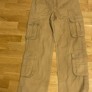 Beiga cargopants med stora fickor - Säljer ett par beiga cargopants med flera stora fickor på benen. Byxorna har en loose passform och är tillverkade i bomull, vilket gör dem både sköna och praktiska. Perfekta för dig som gillar streetwear och vill ha plats för allt du behöver.