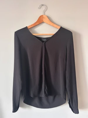 Svart blus från VILA i storlek M - Svart blus från VILA i storlek M med långärmade ärmar och knappdetalj vid mudden. Blusen har en lätt struktur i tyget och en enkel, stilren design med rundad hals och diskret veck framtill. Perfekt för dig som gillar minimalistisk stil.