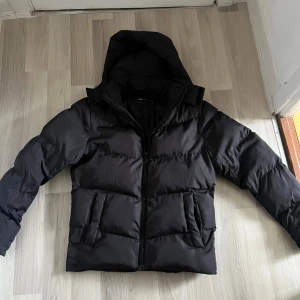 Svart pufferjacka med huva - Svart pufferjacka med avtagbar huva och hög krage. Jackan har två stora fickor framtill och stängs med dragkedja. Den är quiltad och har en clean, modern look som passar perfekt när det är kallt ute.