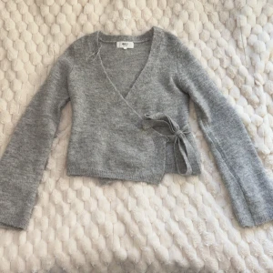 Grå omlottkofta från Gina Tricot - Supermysig grå omlottkofta från Gina Tricot i ullblandning. Koftan har långa ärmar, v-ringad hals och knyt i sidan för en snygg passform. Perfekt att slänga på sig när det är lite kyligt ute. Stilren och enkel att matcha med dina favoritjeans.