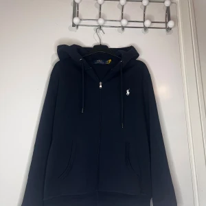 Mörkblå hoodie från Polo Ralph Lauren - Snygg mörkblå hoodie från Polo Ralph Lauren med dragkedja och huva. Tröjan har snörning vid huvan, två stora fickor fram och den klassiska vita Polo-loggan på bröstet. Perfekt för chill dagar och enkel att matcha med jeans eller joggers.